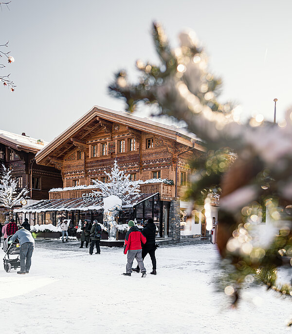 Menschen spazieren bei schönem Wetter auf der schneebedeckten Promenade in Gstaad. Im Vordergrund Äste, im Hintergrund stehen Häuser.