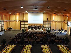 Une grande salle de fête avec quatre rangées de tables dressées et des sièges se trouve au premier plan, plusieurs personnes sont assises sur des chaises au centre, tandis qu’en arrière-plan, une fanfare joue sur scène.
