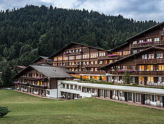 Das grosse Hotel Huus mit vielen Fenstern steht vor einer Rasenfläche, daneben ein Wald von Bäumen auf einem Hügel.