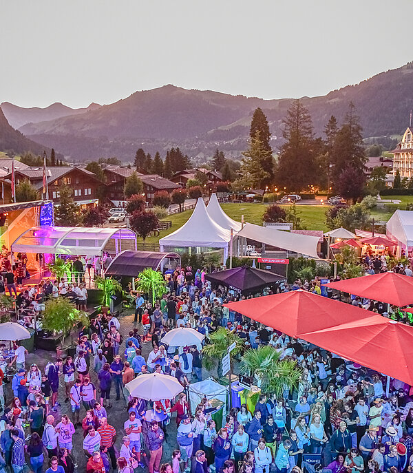 Une place avec des tentes colorées, une scène et beaucoup de monde, avec le village de Gstaad en arrière-plan.