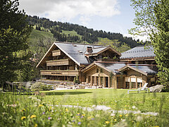 Das Hotel ist in der Bildmitte zu sehen davor ist eine grüne Wiese mit Blumen, rechts und links am Bildrand sind Bäume.