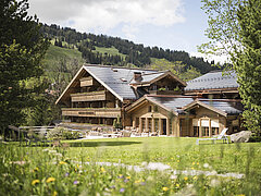 The hotel can be seen in the center of the picture, in front of it is a green meadow with flowers, to the right and left at the edge of the picture are trees.