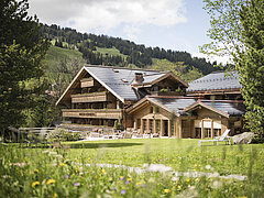Das Hotel ist in der Bildmitte zu sehen davor ist eine grüne Wiese mit Blumen, rechts und links am Bildrand sind Bäume.
