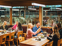 Seven guests sit at two set tables in Michel's Stallbeizli, eating cheese fondue and toasting with glasses of white wine. In the background, you can watch the cows in the barn.