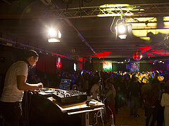  Lots of people dancing in a large hall. In the foreground, a male DJ stands at a mixing deck.