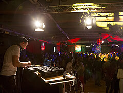 Viele Leute tanzen in einer grossen Halle. Im Vordergrund ein männlicher DJ an einem Mischpult.