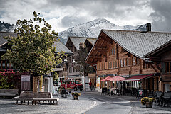 Saanen village centre with typical old chalets, benches, a tree in the foreground and snow-covered mountains in the background.