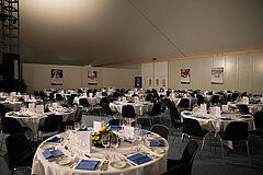 Weisser Raum mit mehreren runden Banketttischen, gedeckt mit weissen Tischtüchern, blauen Servietten, weissen Tellern und Besteck. Pro Tisch acht schwarze Stühle.