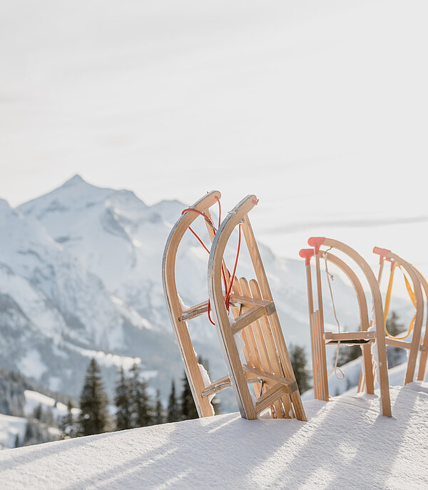 Drei hölzerne Schlitten stehen zuoberst auf dem Berg im frischen Pulverschnee. Im Hintergrund weisse Berggipfel.