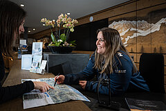 An employee at Gstaad Saanenland Tourism showing a female visitor something on a panoramic map. The counters of the Gstaad tourist office can be seen in the background.