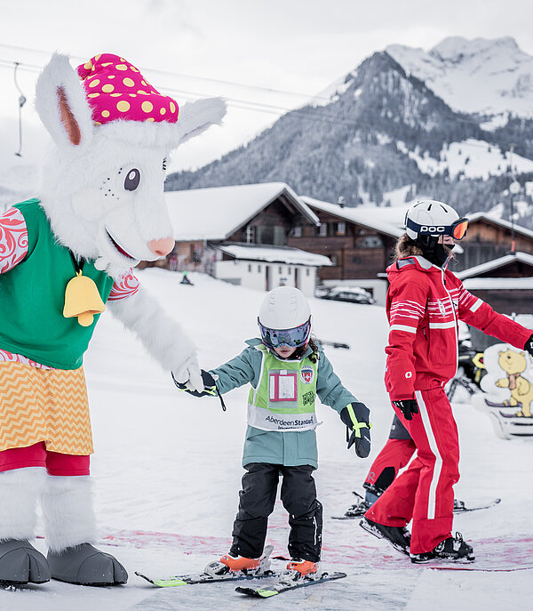 Das Maskottchen Saani im Kinderland der Skischule Gstaad mit einer rot gekleideten Skilehrerin und zwei kleinen Kindern auf Ski.