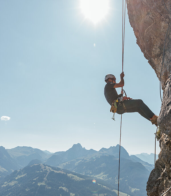 Ein Kletterer seilt sich an einem Seil den Fels hinab, im Hintergrund die Bergkulisse vom Saanenland unter blauem Himmel.