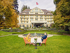 A man and woman sitting at a table on the green lawn in front of the Hotel Le Grand Bellevue.