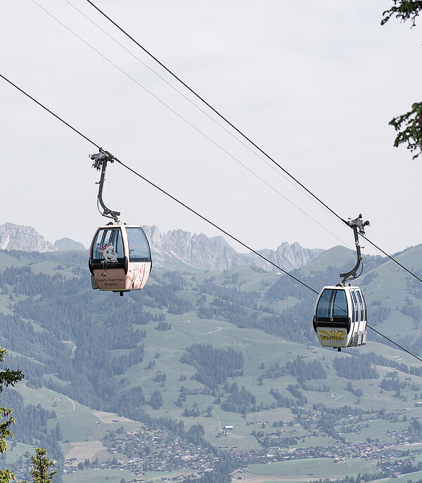 Zwei Gondel welche einen Berg hochfahren. Im Hintergrund sieht man ein Bergpanorama.