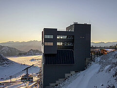 Haut bâtiment sombre. À gauche, une cabine de téléphérique et un télésiège. A droite, un chalet en bois et un pont. En arrière-plan, des montagnes enneigées et le glacier.