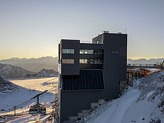 Dunkles hohes Gebäude. Links eine Luftseilbahnkabine und eine Sesselbahn. Rechts ein Holzchalet und eine Brücke. Im Hintergrund verschneite Berge und der Gletscher.