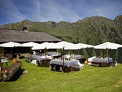 Un groupe de tables recouvertes de nappes blanches et protégées par des parasols dans un pré, avec en arrière-plan une chaîne de montagnes sur fond de ciel bleu.