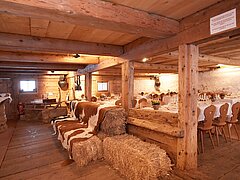 Des tables recouvertes de nappes blanches, des chaises en bois, des bottes de paille et une luge à cornes ainsi que des verres sont disposés dans l’étable rustique d’un chalet d’alpage.