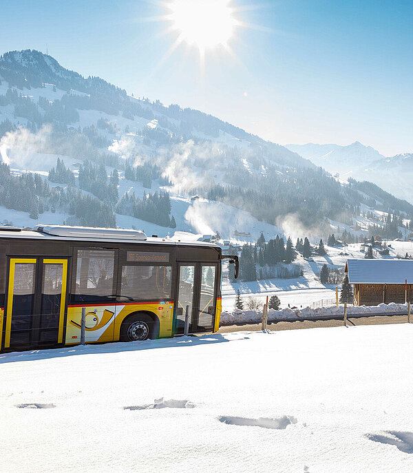 Ein Postauto fährt durch eine Winterlandschaft. Die Sonne scheint. Man sieht auf Gstaad.