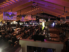 Large hall with lots of people sitting or standing at tables and benches. In the background, a band is playing on a stage. Various American flags with country music motifs hang from the ceiling.