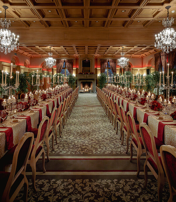 Dans une salle luxueuse, une longue table est dressée avec des bougies, des verres à vin et des fleurs.