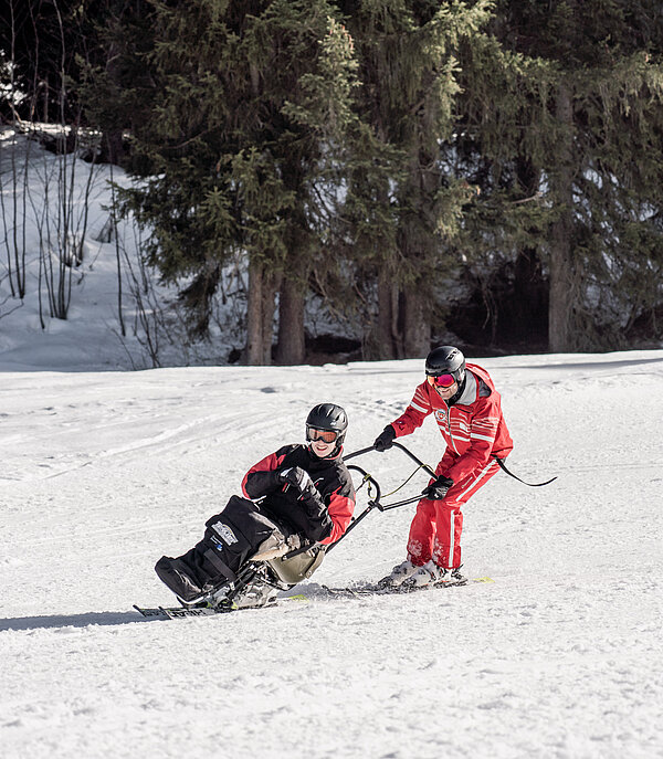Ein Skilehrer in roter Skilehrerbekleidung führte eine Person in einem Sitzski auf der Piste