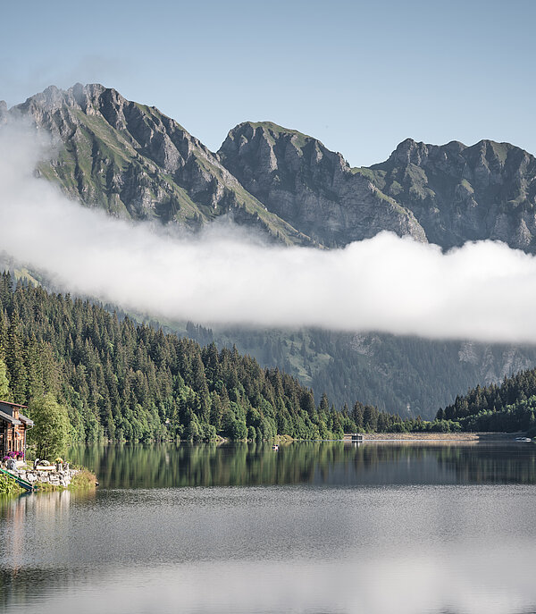 Blick auf den Arnensee mit ruhigem Wasser, einer Alphütte am Ufer, dichten Wäldern und Bergen im Hintergrund, teilweise von Wolken umhüllt.